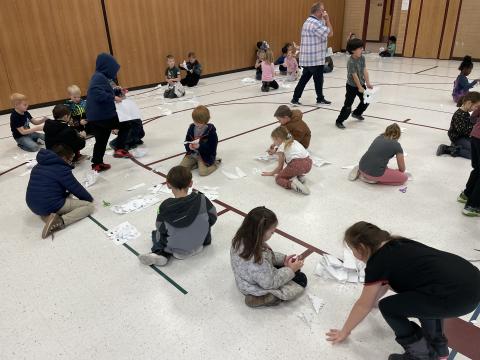 Second grade boys and girls sitting on the gym floor creating their own paper snowflakes
