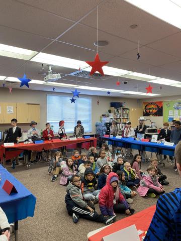 Younger Rees students sitting on the floor listening to 5th grade students present during the wax museum.
