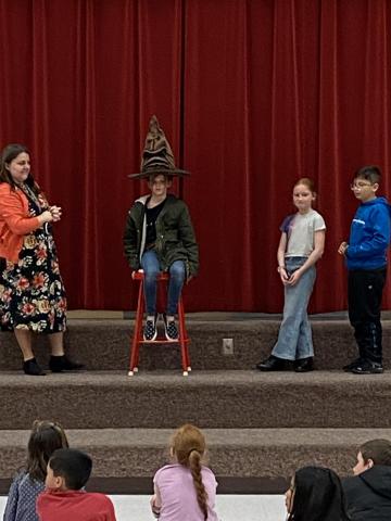 4th grade boy sitting on a stool with soring hat on, girl and boy standing on line beside them. 
