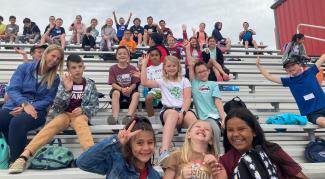 Rees 4th grade students and a technicians smiling as they wait on the bleachers